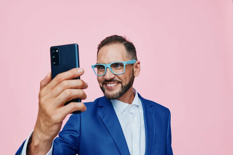 A professional businessman smiles while taking a selfie against a bright colored backdrop, projecting confidence, joy, and approachability in a clean corporate scene.