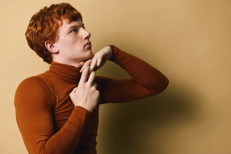 Young man with red hair wearing brown turtleneck poses thoughtfully against beige background in studio. Side profile with hands near neck shows calm and focus.