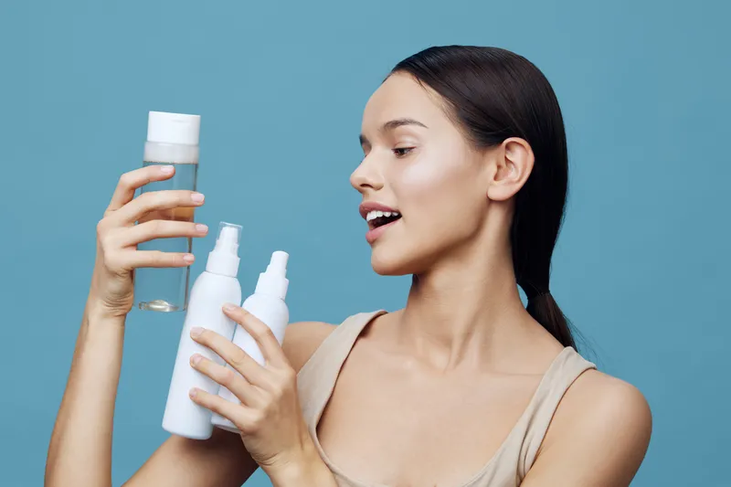 Young woman examining skincare products in minimalist style, expressing curiosity and joy against a soft blue background