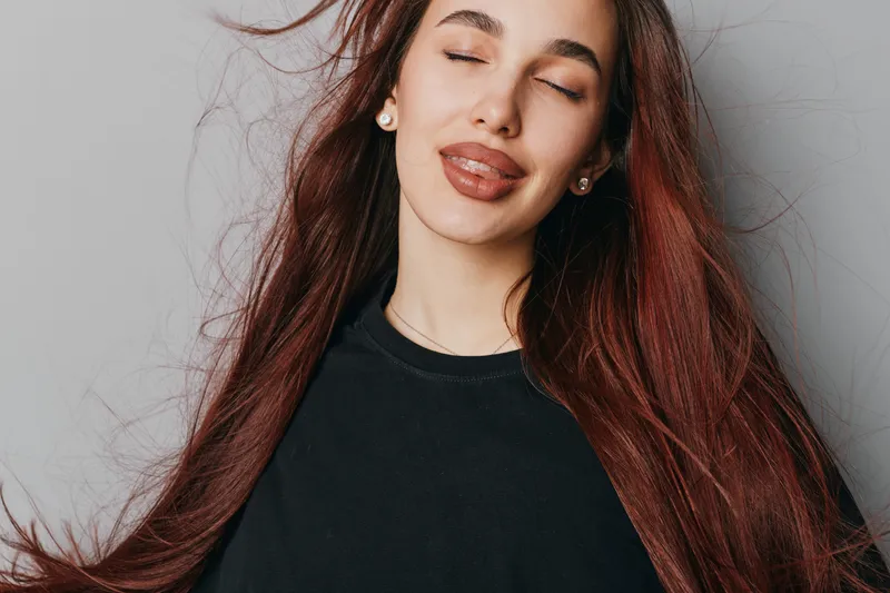 Young woman with long red hair and closed eyes making playful face with tongue out wearing black top in studio. Beauty, fun and carefree mood with natural expression on gray background.