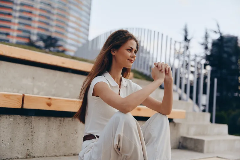 Young woman sitting on urban stairs smiling thoughtfully in casual white outfit during daytime outdoor lifestyle scene KEYWORDS: young woman, casual outfit, sitting outdoors, smiling, thoughtful expre