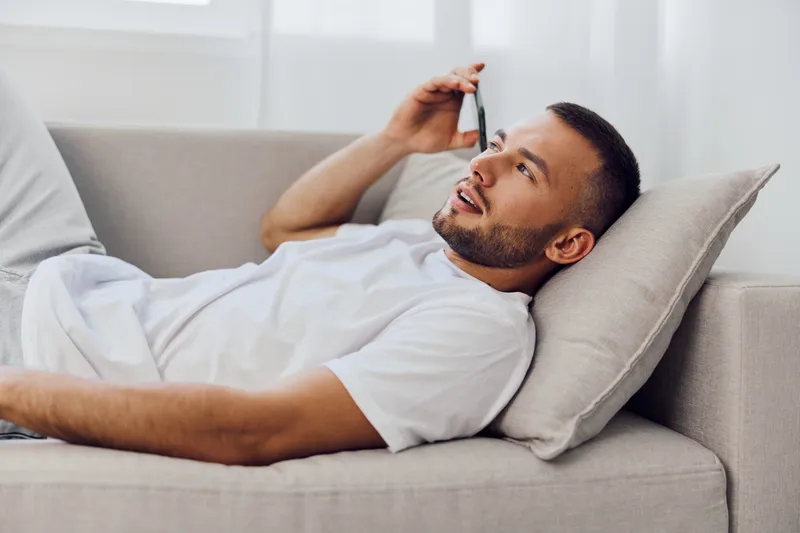 Relaxed man on the couch enjoying an authentic phone call, embodying soft masculinity and emotional connections. A moment of genuine vulnerability in a cozy home setting.