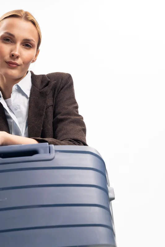 Young woman in brown jacket holding blue suitcase looks confidently at camera. Studio portrait on white background. Concept of travel, business, and modern lifestyle.