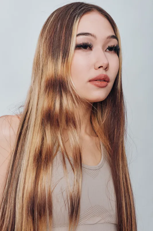 Young woman with long straight hair and natural makeup posing on neutral background, looking away thoughtfully in casual sleeveless top portrait.