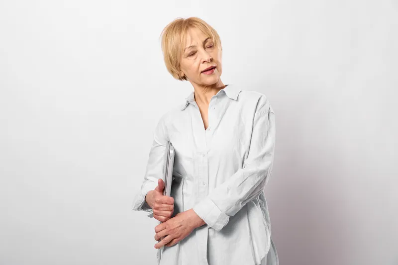 Senior woman in healthcare context holds her aching arm, looking concerned, Studio background emphasizes arthritis pain, tenderness, discomfort, stress, and wellness