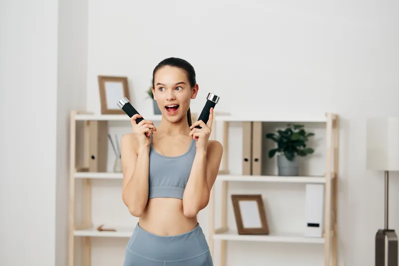 Woman fitness enthusiast in a bright home gym, smiling as she lifts dumbbells for a full body workout, showcasing energy, motivation and an active lifestyle in a comfortable living space.