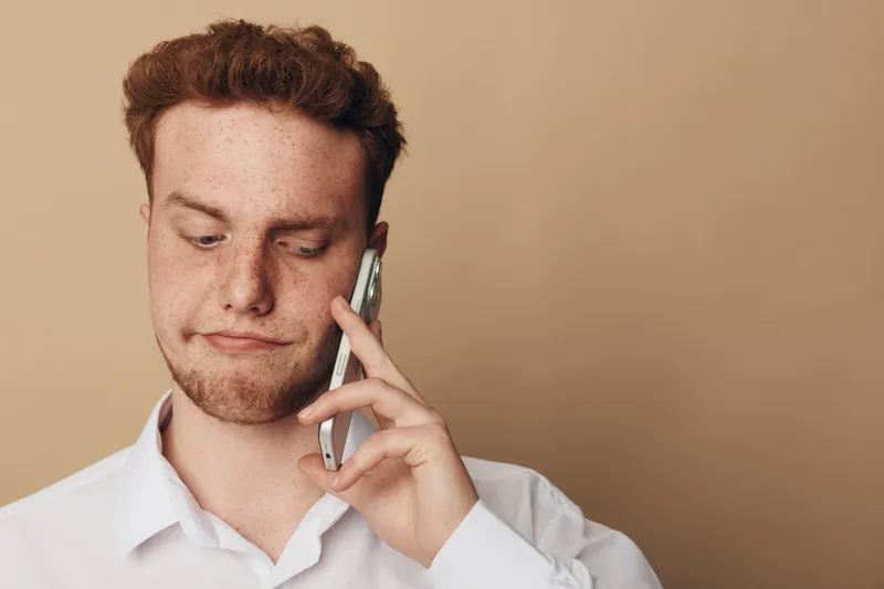 Young man with red hair and freckles wearing white shirt talks on smartphone with doubtful expression on beige background. Concept of communication, technology, and uncertainty.