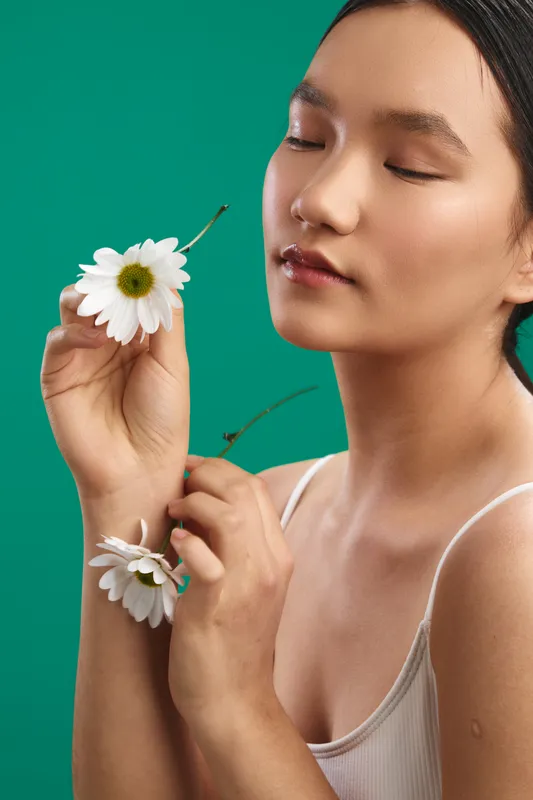 Asian woman holds white daisies against a teal backdrop, eyes closed and serene, conveying calm beauty and mindful simplicity in a studio portrait.
