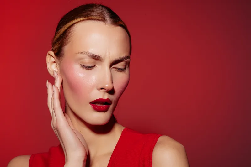 Woman with red lipstick and red dress poses with closed eyes and hand near face on red background. Concept of beauty, fashion, makeup, and elegance in studio lighting.