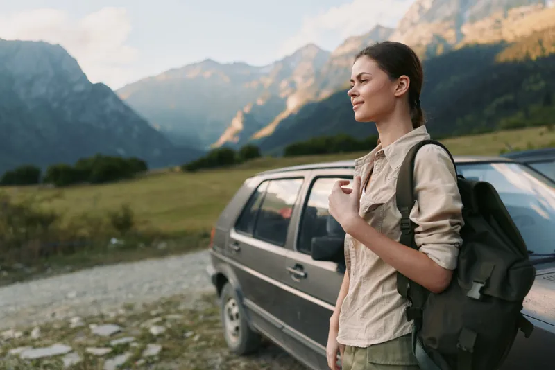 Young woman with a backpack stands beside a car in a mountain valley, ready for a hike and enjoying the peaceful outdoor scene, sunrise glow highlighting rugged peaks.
