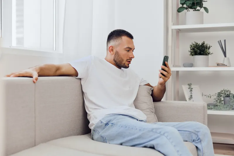Relaxed man using smartphone on a cozy couch in a bright living room, highlighting modern minimalism and tranquility with a touch of contemporary style.
