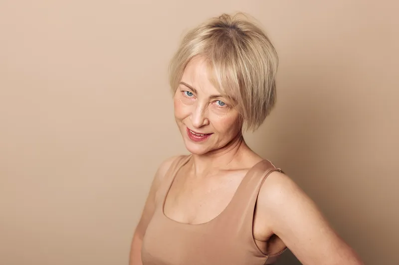 Smiling mature woman with short blonde hair poses confidently against beige background. Natural beauty portrait of joyful middle aged female with clear skin and casual outfit. Concept of aging grace.