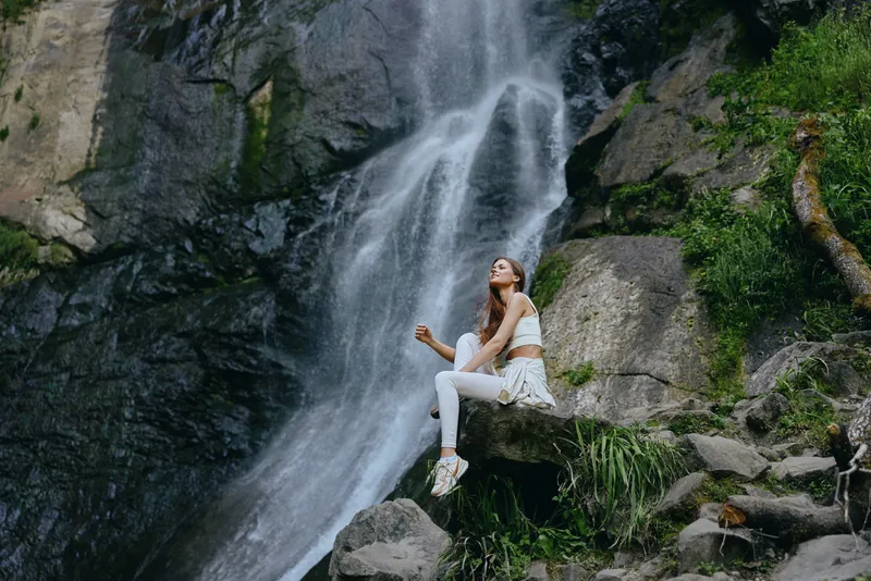 Woman meditating near waterfall dressed in white activewear sitting peacefully on rocks surrounded by lush greenery and natural scenery in outdoor environment.