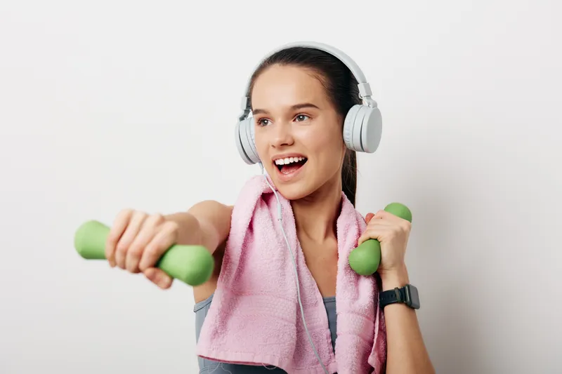 Woman fitness enthusiast wearing headphones, towel around neck, and light dumbbells in hand, mid workout in a bright studio, conveying energy, discipline, and healthy living.
