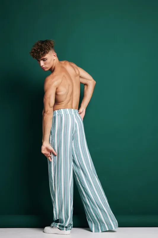 Male model with curly hair, shirtless pose, striped loose pants and sneakers standing against dark green background for fashion and style concept at studio.