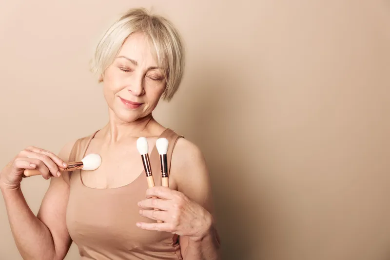 Smiling mature woman with short blonde hair applies makeup brushes to chest and face on beige background. Concept of beauty routine, self-care, and natural skincare for older women.