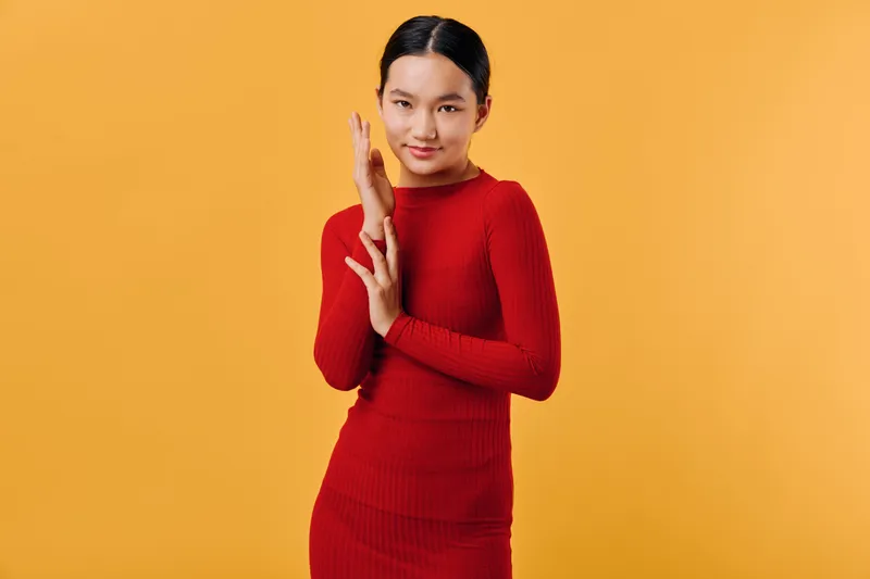Young woman in red dress posing with hand gestures on orange background, confident model with natural makeup and neat hairstyle in studio photo shoot session.