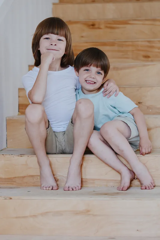 Two boys sitting on stairs, smiling and enjoying togetherness, playfully posing in casual clothing, reflecting warmth and joy in a cozy setting.