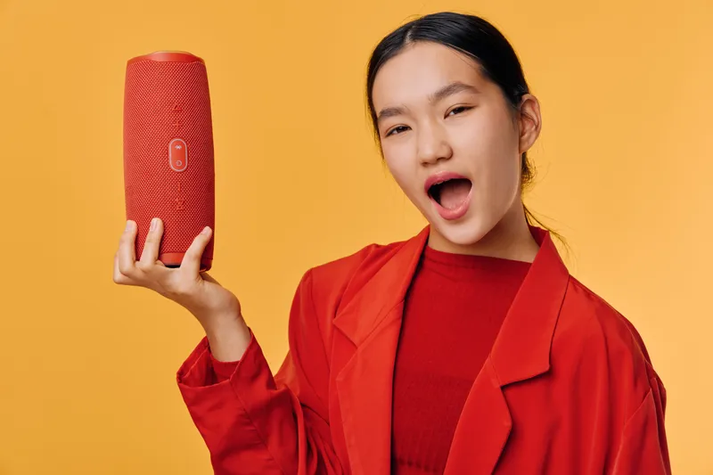 Young woman with black hair wearing red jacket and shirt holds red wireless speaker while winking and smiling against orange background for technology and music concept.