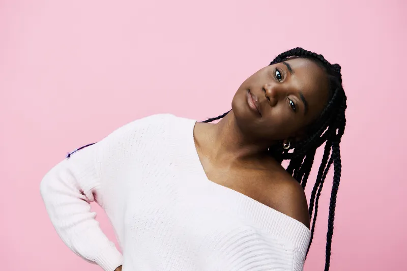 Young black woman with braided hair in an off shoulder sweater against a pink background, conveying confidence and casual elegance