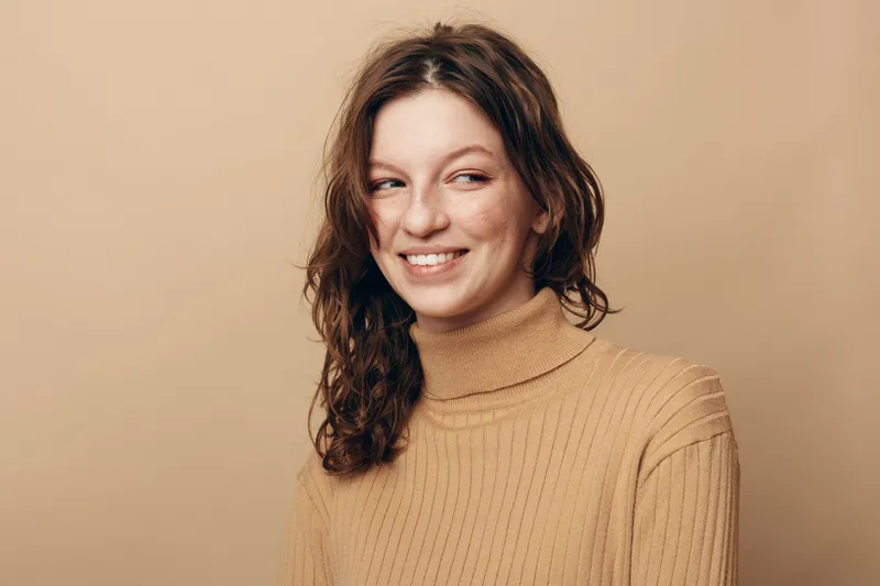 Smiling woman with curly hair wearing beige turtleneck sweater looks aside on beige background. Portrait of happy young adult female with natural makeup and casual style in warm studio lighting.