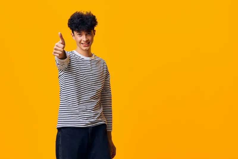 smiling man with thumbs up wearing striped long sleeve shirt and dark pants standing against bright orange background expressing positivity and confidence