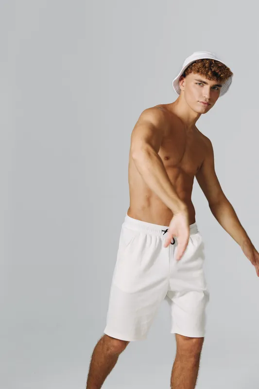 Young man with curly hair wearing white shorts and bucket hat posing shirtless against gray background. Casual style, summer fashion and confident male model in studio lighting.