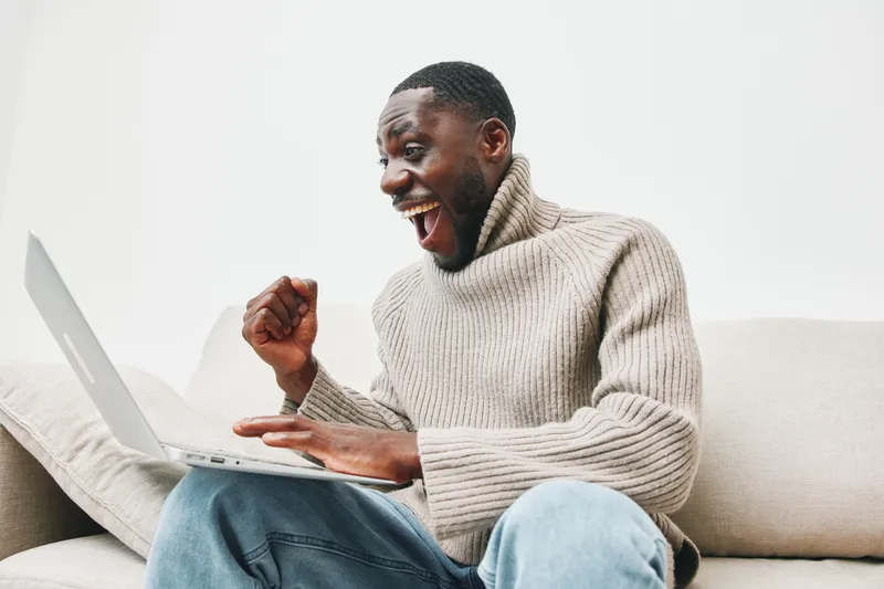 Happy Black man celebrating success on laptop in cozy sweater, expressing joy and excitement while sitting on couch, modern home interior design