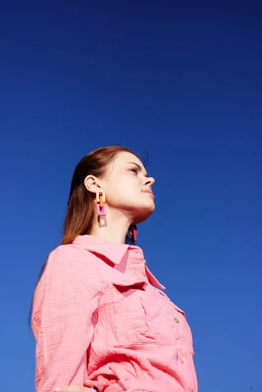 Young woman in bright pink attire with earrings stands against a clear blue sky, exuding confidence and positivity during sunny weather