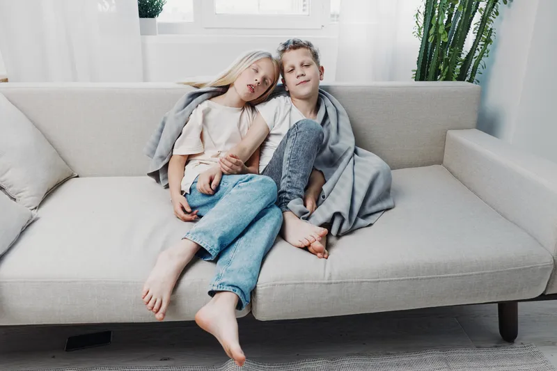 Two children relax barefoot on a beige sofa in a cozy bright living room. They share a gray blanket, showing sibling comfort and casual family moments at home.