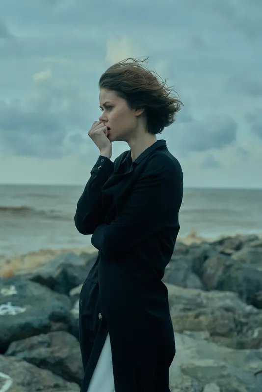 Windy seaside scene with a contemplative woman in a dark coat standing on a rocky shore, moody landscape and ocean beyond