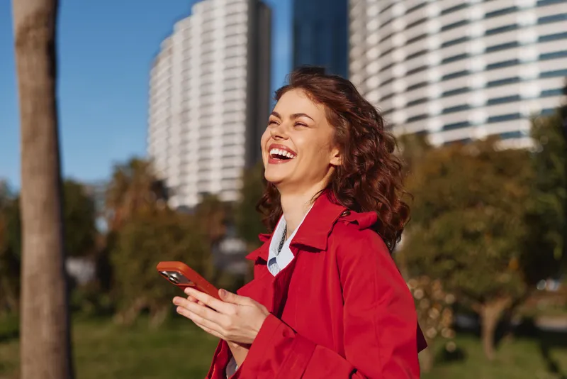 A happy young woman in a red coat laughs while holding a smartphone outdoors with modern buildings and trees in the background on a sunny day.