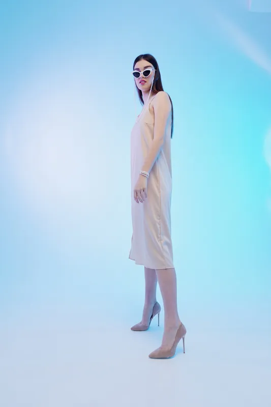 Slim woman in a beige dress and high heels poses confidently wearing sunglasses. Studio shot with soft blue lighting. Concept of woman beauty fashion and elegance.