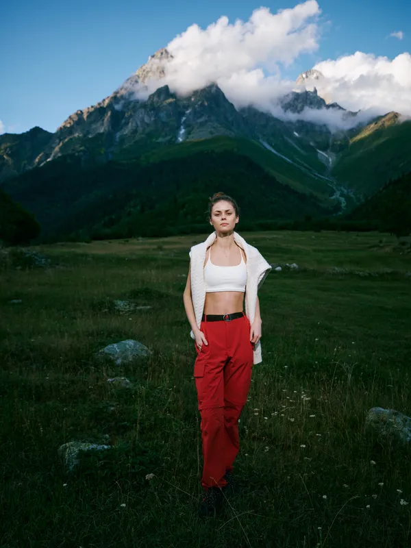 Mountain landscape with a stylish woman standing in a meadow, cropped top and red pants, light jacket draped over shoulders, dramatic peaks in the distance, serene outdoor fashion in nature