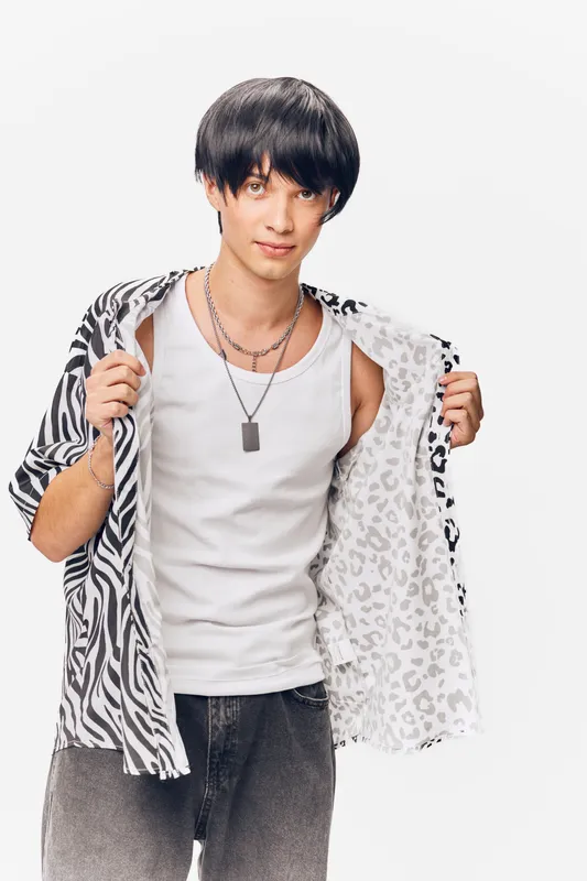 Young man wearing a black and white patterned shirt over a white tank top, posing with a trendy hairstyle and necklace, isolated on white background.