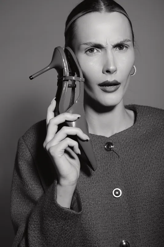Caucasian woman with light skin holds high heel shoe to ear as if it is a phone, wearing textured coat. Concept of confusion and creativity.