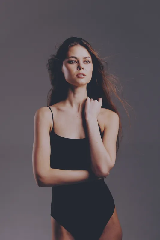 Elegant woman posing confidently in black bodysuit with neutral background, natural lighting, and soft expression