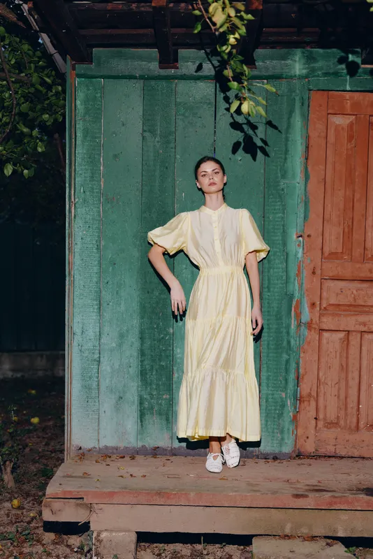 A stylish woman stands on a wooden porch wearing a soft yellow dress, striking a confident pose against a green wall and orange door, creating a cinematic fashion moment