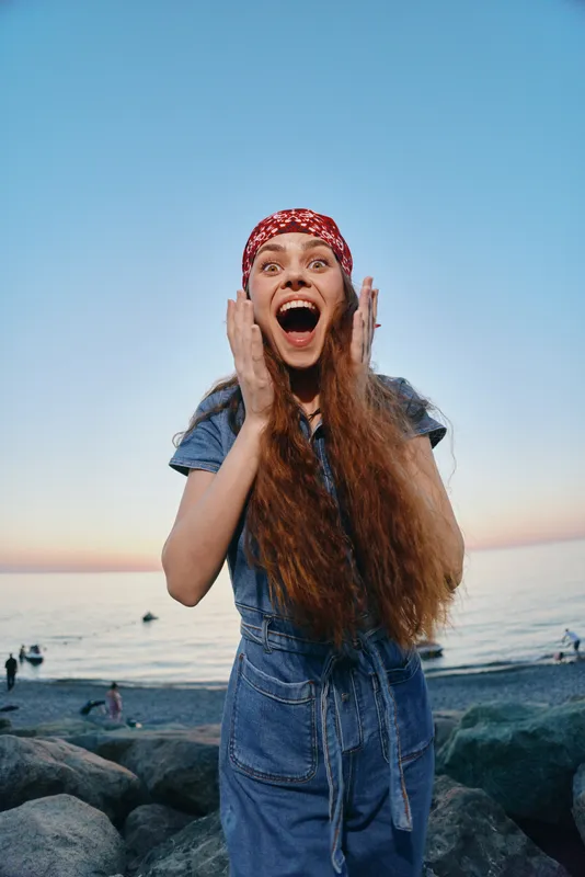 Excited woman in modern boho-western denim street style with a red bandana smiles joyfully by the seaside during golden hour, film color enhancing warm tones.