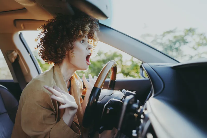 Young woman with curly hair drives a car, mouth open in surprise as she reacts to something off screen, sunlight fills the cabin and highlights her beige jacket.
