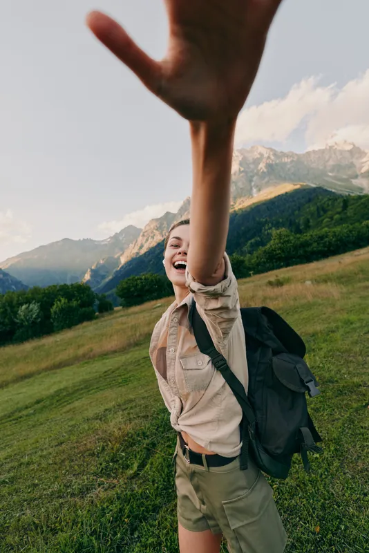 A young woman hiker with a backpack reaches toward the camera in a sunlit mountain meadow, capturing a spontaneous moment of adventure and confidence.