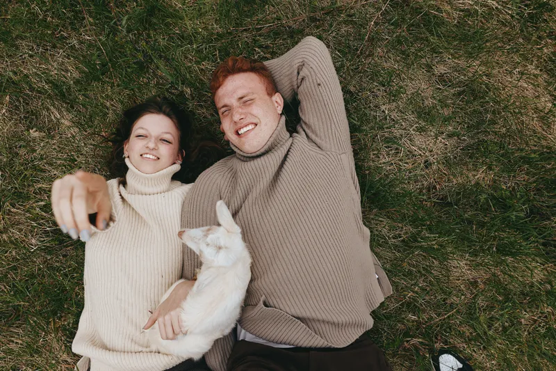 A smiling couple lies on the grass in knit sweaters, with a playful goat kid beside them. A lifestyle scene that highlights outdoor leisure, warmth, affection, and the bond between humans and animals.