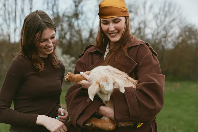 Two women outdoors interact with a fluffy goat kid in a rural farm setting, a casual lifestyle moment that highlights care, friendship, nature, and the simple charm of farm life on a bright day.