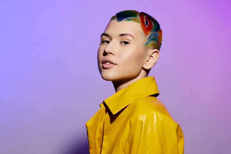 young woman with colorful rainbow hairstyle wearing bright yellow coat stands confidently against purple background, showcasing unique creative fashion and self-expression.