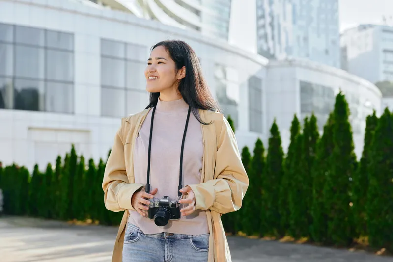 Camera in hand, woman smiles while exploring an urban campus with green hedges and glass buildings, capturing travel, discovery and everyday creativity in outdoor photography.