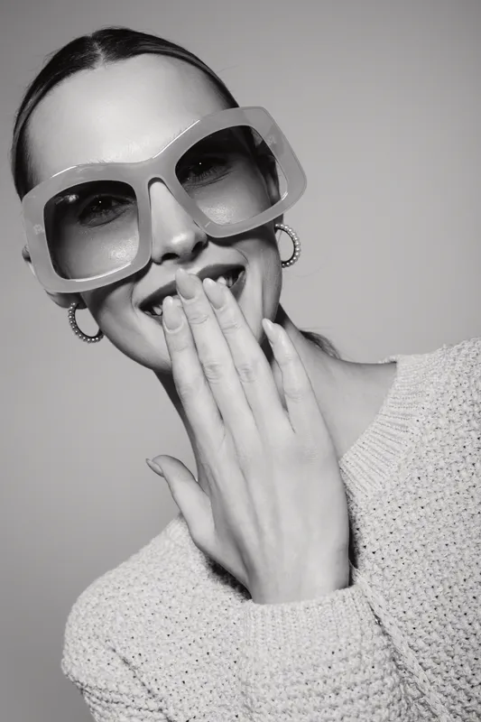Happy Caucasian woman with light skin wearing large glasses laughs covering mouth with hand in studio. Concept of joy, fashion, and candid moment.