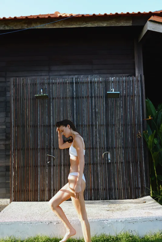 Young woman in white swimsuit walks barefoot past outdoor bamboo shower near wooden cabin on a sunny day, enjoying a relaxed lifestyle moment in nature.