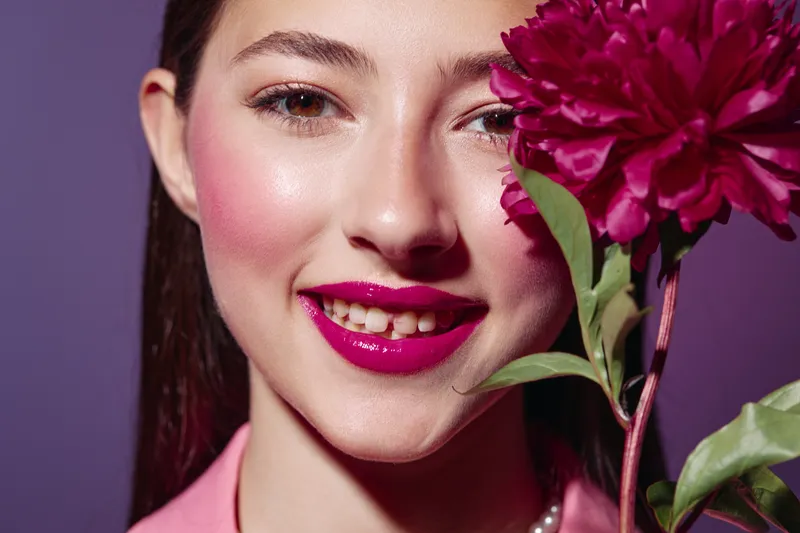 Close-up of smiling young woman with vibrant pink lipstick and flushed cheeks holding a red flower near her face on purple background. Beauty creative fashion concept.