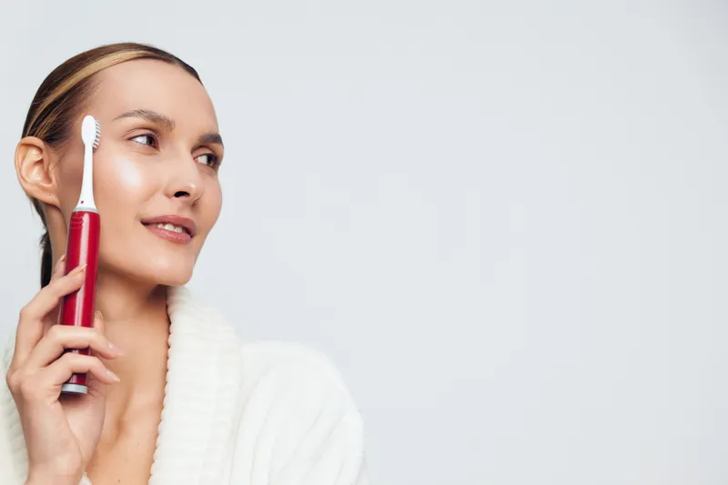 A young woman in a white bathrobe holds a red electric toothbrush near her face against a plain background. Concept for dental care, oral hygiene, and personal grooming.