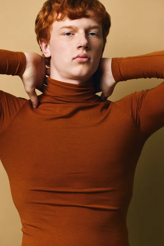 Young man with red hair and serious expression posing with hands behind head wearing brown turtleneck sweater in front of plain background indoors studio portrait.